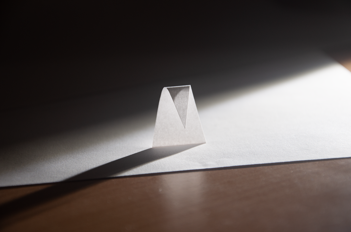 photo depicting abstract sadness - bent paper triangle in dark with light from behind creating a long shadow 